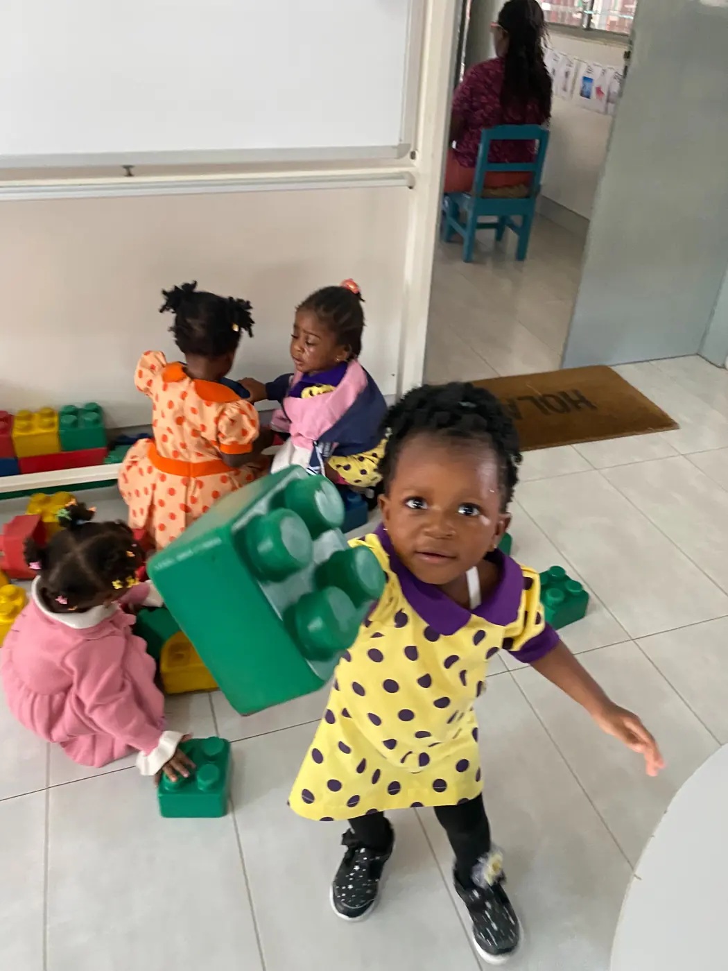 A toddler carrying a big green building block while friends play